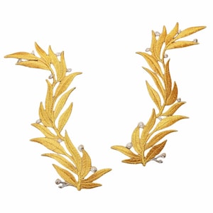 Puede incluir: Dos coronas de laurel doradas con cuentas blancas, cada una con una forma curva y detalles intrincados.