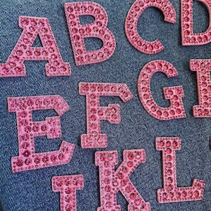 May include: Pink jeweled letters of the alphabet A, B, C, D, E, F, G, I, K, and L on a blue denim background.