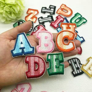 Puede incluir: Una colección de parches de letras bordadas en tela de varios colores. Las letras A, B, C, D y E están enfocadas, con tonos azules, rosas, naranjas, rojos y verdes. Otras letras están dispersas en el fondo, sobre un fondo blanco.