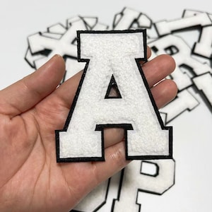 Puede incluir: Letra A de fieltro blanco con contorno negro. La letra es gruesa y tiene un estilo universitario clásico.