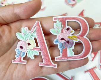 Könnte beinhalten: Zwei rosa Filzbuchstaben, A und B, mit weißen Stickereien und Blumendesigns. Die Buchstaben werden in einer Hand gehalten.