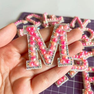May include: Pink and white letter M made of rhinestones and pearls. The letter is on a white background.