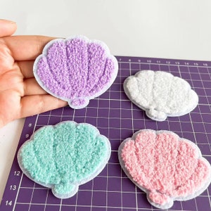 Può includere: Quattro toppe di tessuto peloso a forma di conchiglia in diversi colori: viola, bianco, azzurro e rosa. Le toppe sono su uno sfondo a griglia viola.