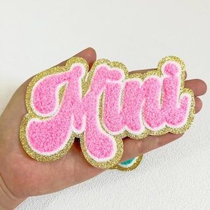 May include: Pink and gold felt letter patch with the name "Kimi" in a retro style font.