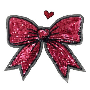 Puede incluir: Un gran lazo rojo con lentejuelas, contorno negro y un pequeño corazón rojo encima. El lazo es de un tono rojo vibrante, y las lentejuelas le dan un aspecto brillante. El diseño está sobre un fondo blanco.