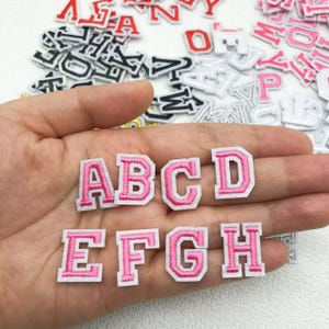 Może przedstawiać: Haftowane litery z różowej tkaniny z białymi obwódkami. Widoczne litery A, B, C, D, E, F, G i H, trzymane w dłoni. Inne litery w różnych kolorach są rozrzucone w tle.