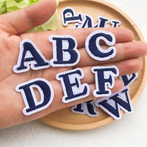May include: A selection of navy blue embroidered fabric letters, including A, B, C, D, E, F, and W, with white borders. The letters are presented on a wooden tray, suitable for crafting or customisation.