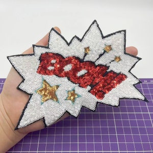 Può includere: Una toppa di paillettes bianche e rosse con la parola "BOOM" in un font in stile fumetto. La toppa è circondata da paillettes bianche e paillettes dorate a forma di stella.