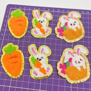 May include: Six felt Easter-themed patches with gold glitter trim. Two patches feature a white bunny holding a carrot, two patches feature a white bunny in a basket with a pink bow, and two patches feature a carrot.