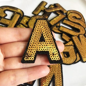 Puede incluir: Un conjunto de letras del alfabeto de lentejuelas doradas, cada letra está recortada individualmente y respaldada con fieltro negro. Las letras están dispuestas en una pila, con la letra A en la parte superior.