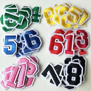 May include: A collection of embroidered fabric numbers in colours such as green, yellow, blue, red, pink, and black. Each number has a white border and a geometric shape. These numbers are suitable for patches or appliques.