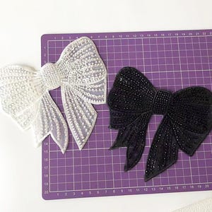 May include: Two fabric bows, one white and one black, with a beaded design. The bows are on a purple gridded surface.