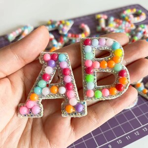 May include: Two colourful letters, A and B, made of white felt with rainbow coloured beads and rhinestones around the edges.