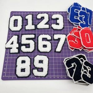 以下が含まれることがあります： 0から9までの白いフェルトの数字のセットで、黒いアウトラインが付いています。数字は紫色のカッティングマットに2行に並んでいます。背景には、赤、青、黒のフェルトの数字のセットもあります。