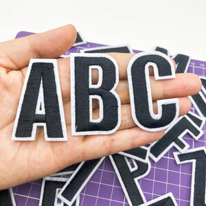 Peut inclure: Lettres en feutre noir avec des contours blancs, épellant "ABC". Les lettres sont disposées en rangée et sont tenues dans une main. Les lettres sont sur un fond de grille violet.