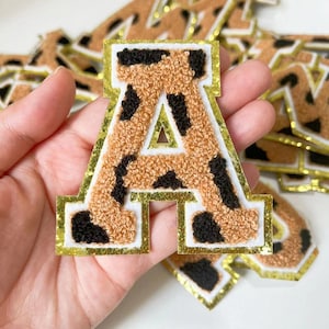 Può includere: Un patch a forma di lettera A, peloso, con bordo dorato e un motivo leopardato marrone e nero.
