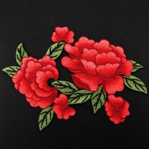 以下が含まれることがあります： 緑の葉が付いた赤い刺繍の花のアップリケ。花は房状に集まっており、繊細で重ねられたデザインになっています。