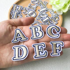 Könnte beinhalten: Gestickte Buchstaben-Patches in Beige und Marineblau. Die Buchstaben A, B, C, D, E und F sind sichtbar, weitere im Hintergrund. Ideal zum Personalisieren von Kleidung, Taschen oder anderen Stoffartikeln.