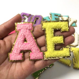 May include: Pink and yellow chenille letter patches with gold glitter borders. The letter "A" is pink, and the letter "E" is yellow. Other colorful letters are visible in the background.