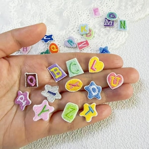Puede incluir: Un conjunto de parches bordados con letras del alfabeto, en diversas formas como estrellas y corazones. Cada parche presenta una letra diferente en un color contrastante, ideal para manualidades y personalización.