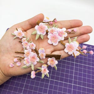 May include: Five pink embroidered flower patches with brown stems and green leaves. The patches are arranged on a hand, with the background being a purple grid.