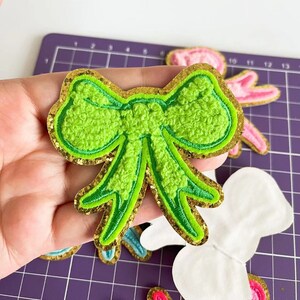 May include: A green fuzzy bow with a gold glitter outline. The bow is a fabric patch with a stitched edge.
