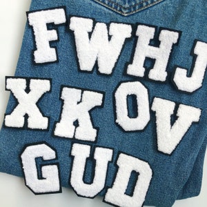 Puede incluir: Letras blancas de fieltro con contornos negros sobre un fondo de mezclilla azul. Las letras deletrean "FWHJ XKOV GUD".