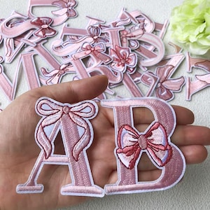 May include: Pink embroidered letter patches with bow accents. The letters "A" and "B" are held in a hand, with other letters scattered in the background. The patches are a light pink colour with white and darker pink details.