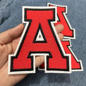 Può includere: Lettera A in feltro rosso con contorno nero e retro bianco. La lettera è grande e ha uno stile varsity classico.