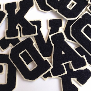 Könnte beinhalten: Eine Ansammlung schwarzer Filzbuchstaben mit cremefarbenem Rand. Die Buchstaben in einer klassischen College-Schrift sind auf einer weißen Oberfläche verstreut. Ideal für Bastel- oder Personalisierungsprojekte.