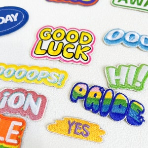 以下が含まれることがあります： さまざまなフレーズが刺繍されたカラフルなパッチのコレクション。「GOOD LUCK」、「PRIDE」、「YES」、「HI！」の文字が含まれています。パッチはさまざまな形とサイズで、明るい色と遊び心のあるフォントが特徴です。