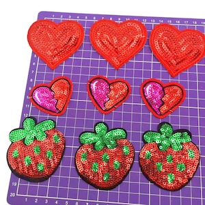 May include: Nine sequined patches in the shape of hearts and strawberries. The hearts are red, with some split in half, showing pink sequins. The strawberries are red with green leaves and green sequin seeds. The patches are on a purple cutting mat.