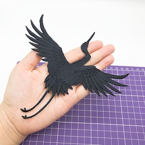 Może przedstawiać: Czarna, haftowana naszywka z żurawiem z rozłożonymi skrzydłami, szczegółowymi piórami i długimi nogami. Naszywka jest elementem dekoracyjnym, prawdopodobnie do odzieży lub akcesoriów, i jest trzymana w dłoni na białym tle.