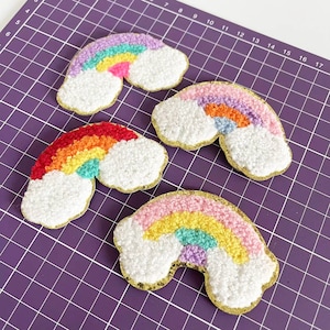 Peut inclure: Quatre patchs arc-en-ciel et nuage avec une texture moelleuse et une bordure dorée pailletée. Les arcs-en-ciel sont faits de fils de différentes couleurs et les nuages sont blancs.