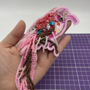 May include: Pink and red beaded bird applique with brown and white beads, rhinestones and sequins. The bird has a long tail with pink, brown and white beads.