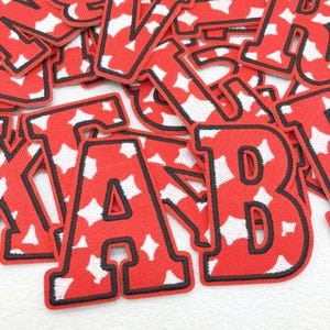 Puede incluir: Un conjunto de parches de letras de tela roja con un patrón de estrellas blancas. Cada letra tiene un borde negro. Las letras están en varias fuentes y tamaños, adecuadas para proyectos de manualidades o personalización.