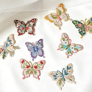 May include: A collection of eight embroidered butterfly patches featuring intricate floral and botanical patterns in various colors including red, blue, purple, yellow, green, and pink. Each patch displays detailed thread work and distinct wing shapes.
