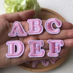 May include: Pink embroidered fabric letters A, B, C, D, E, and F with a white border. The letters are in a serif font and are displayed on a wooden plate. The letters are a light pink colour.