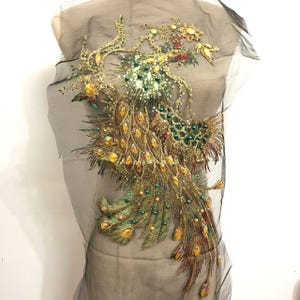 Puede incluir: Una aplicación bordada de pavo real dorada y verde con lentejuelas y cuentas. La aplicación está sobre una tela de malla negra.