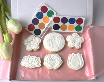Può includere: Una scatola regalo piena di biscotti di zucchero bianchi decorati con vari motivi floreali e le parole "Happy Mother's Day". La scatola contiene anche due set di acquerelli, uno con i colori rosso, verde, blu e arancione, e l'altro con i colori viola, rosso, blu, arancione e verde.
