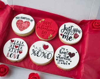 May include: Six round Valentine's Day cookies with various designs and messages. The cookies are decorated with red, black, and white icing. The messages on the cookies include "You Complete Me", "Happy Valentine's Day", "Just You and Me", "XOXO", "My Heart is Wherever You Are", and "Love".