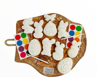 Può includere: Un set di biscotti di zucchero a tema pasquale su un tagliere di legno con vernice e pennelli per la decorazione. I biscotti hanno la forma di conigli, uova e una farfalla. La vernice è in un piccolo contenitore di plastica con una varietà di colori.