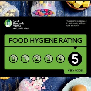 Może przedstawiać: Zielony i czarny znak z tekstem "Food Hygiene Rating" i skalą od 0 do 5 z czarnym k&oacute;łkiem wok&oacute;ł każdej liczby. Liczba 5 jest wyr&oacute;żniona w większym k&oacute;łku, a pod nią znajduje się tekst "Very Good". Znak znajduje się na drewnianej powierzchni z r&oacute;żnymi produktami spożywczymi w tle.