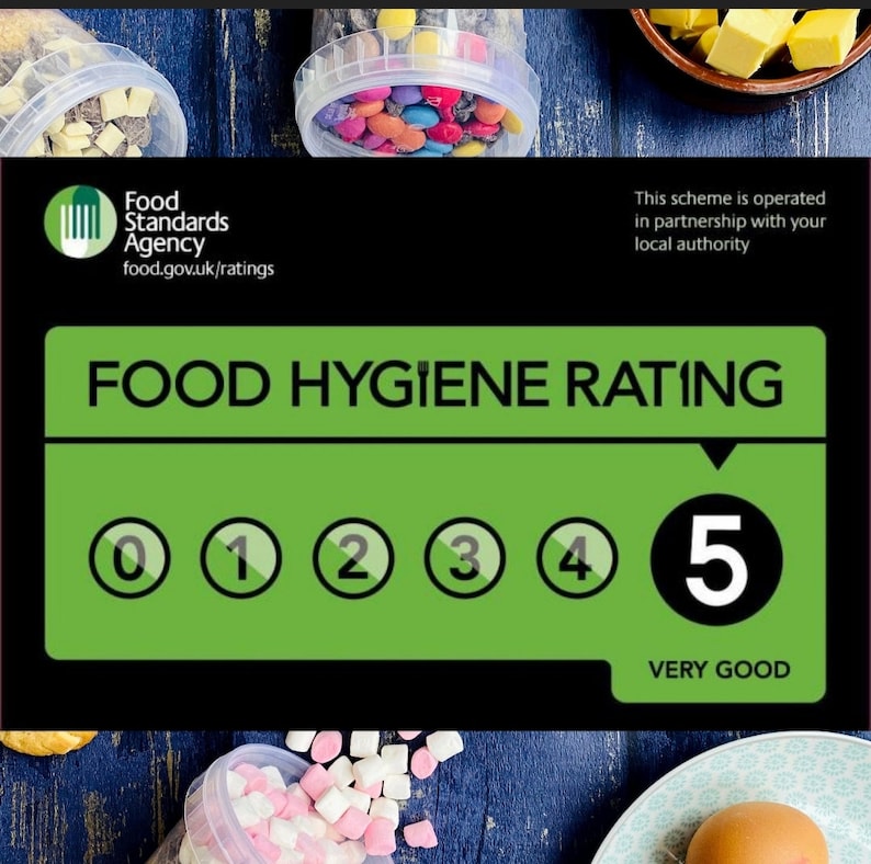 Może przedstawiać: Zielony i czarny znak z tekstem "Food Hygiene Rating" i skalą od 0 do 5 z czarnym k&oacute;łkiem wok&oacute;ł każdej liczby. Liczba 5 jest wyr&oacute;żniona w większym k&oacute;łku, a pod nią znajduje się tekst "Very Good". Znak znajduje się na drewnianej powierzchni z r&oacute;żnymi produktami spożywczymi w tle.