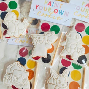 May include: A set of individually wrapped paint-your-own cookie kits, each featuring a white character-shaped cookie, a paint palette card with colourful circles, and a small wooden paintbrush, labelled with the text Paint Your Own Cookie.