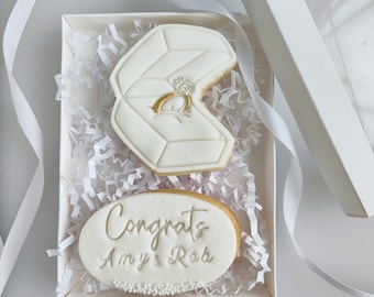 May include: A white gift box with white tissue paper and two white cookies. One cookie is shaped like a ring box with a gold ring inside. The other cookie is oval shaped with the text "Congrats Amy & Rob" in gold lettering.