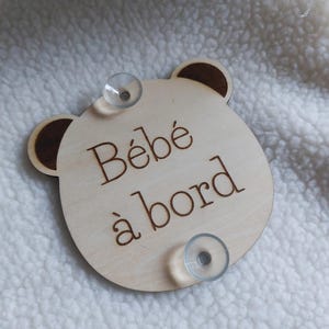 May include: A wooden sign with a bear face and the text "Bébé à bord" in French. The sign has two suction cups on the back.