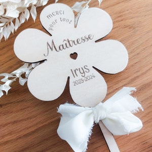 May include: A wooden flower-shaped sign with the text "Merci pour tout Maitresse Irys 2023-2024" and a small heart. The sign is attached to a wooden stick with a white ribbon bow.