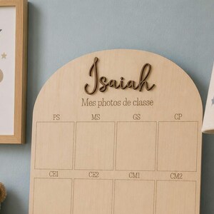 May include: A wooden photo display board with the name "Isaiah" in a cursive font. The board has spaces for school photos labeled PS, MS, GS, CP, CE1, CE2, CM1, and CM2. A framed bear illustration and a lamp with star cutouts are in the background.