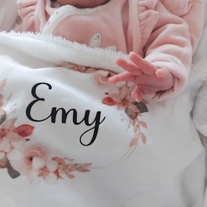 Puede incluir: Una manta de bebé blanca con un diseño floral y el nombre "Emy" impreso en negro. Un bebé está acostado en la manta con un mono rosa con volantes.